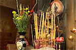 An altar in a Buddhist temple in Vietnam. In the foreground, an urn is filled with burning incense sticks giving off smoke. A flower urn provides some color while a statue with many arms in the background completes the image.