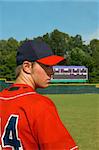 young teen male stands on baseball field in red navy uniform.  He is looking sideways.  Scoreboard is in background of picture.