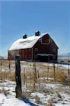 Rustic red wooden barn sits in a field of white.  Pasture has snow, fence and stile.  Snow blows from roof.  Blue skies.