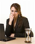 Young brunette woman working at her desk on a laptop computer staring at an old hourglass time while biting her finger with a worried expression