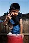 Young asian boy kneeling on top of a red punching bag outside beside a tall wooden fence wearing jeans and black tshirt making hand gesture