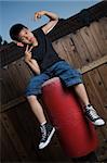 Young asian boy sitting on top of a punching bag outside beside a tall wooden fence smiling wearing jeans and black tshirt making hand signals