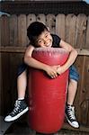 Young asian boy holding on to a punching bag outside beside a tall wooden fence smiling wearing jeans and black tshirt