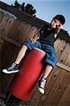 Young asian boy sitting on top of a punching bag outside beside a tall wooden fence smiling wearing jeans and black tshirt