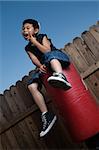Young asian boy sitting on top of a punching bag outside beside a tall wooden fence resting his chin on hand wearing jeans and black tshirt