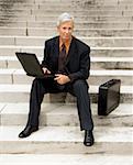 Caucasian middle aged businessman sitting on steps outdoors with laptop and briefcase looking at viewer.