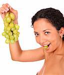 A smiling woman holds a bunch of grape in her hand and eats, standing on white background.