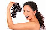 A smiling, happy woman holds a bunch of dark grapes in her hand and eats grape, standing on white background.