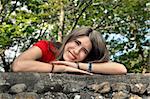 Female teen lays her head on her arms on a stone wall.  Red shirt and a big smile.