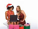 Two attractive females (one ethnic in sexy Mrs. Santa Claus costume and one Caucasian blonde in red dress) sitting surrounded by shopping bags with Christmas presents and browsing internet on notebook portable computer, while holding credit card in hand