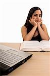 Young Hispanic woman sitting in front of desk with a pile of thick textbooks resting chin on hands with elbows on the table with laptop computer in front