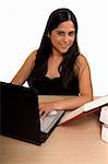 Young Hispanic woman sitting in front of desk reading a textbook and typing on a laptop looking up smiling