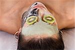 Woman lying down on her back in spa, with special facial mask applied on her face. The mask consist of three colors, each cover different part and eyes are covered by slice of kiwi