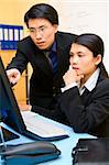 A business man is pointing at the computer screen which his partner works on.