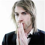 Young man with long, blond hair has a serious look in his eyes. He is holding his hands together as in prayer.