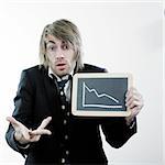 A portrait of a young man in a business suit and tie holding a chalkboard that shows a picture of a graph that is descending. He is making a gesture with his other hand and has a worried expression on his face.