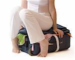 Barefooted woman sitting on a suitcase crammed full of clothes, isolated, with clipping path