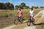 Senior couple bicycling at the beach, wearing safety helmets and sunglasses.