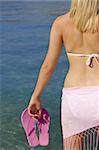 Rear view of a beautiful young blond woman standing by a deep blue sea wearing a white bikini a pink sarong and carrying flip flops