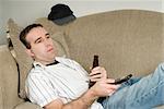 A young man holding a beer while changing channels with a tv remote control