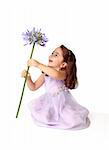 A little girl sitting on the floor in pretty mauve dress decorated with floral appliques, is spinning a large stemmed agapanthus lily flower in bloom, with much delight and excitement.