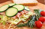 Tasty open sandwich with green lettuce, grated cheese, smoked ham and cucumber on wholewheat bread with rosemary and tomatoes on chopping board