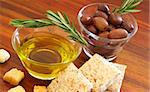 Two jars of black olives with stick of rosemary, olive oil, slices of bread and croutons on wooden table background