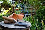 Gardening utensils and flower pots resting on a stool in a green garden with a greenhouse out of focus in the background