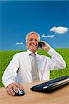 Business concept shot showing an older male executive sitting at a desk, using his computer and talking on his cell phone in a green field with a blue sky complete with fluffy white clouds. Shot on location not in a studio.