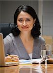 A young businesswoman is sitting at a desk with a computer.  She is smiling at the camera.  Vertically framed shot.