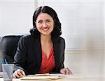A young businesswoman is seated at a desk in an office and is smiling at the camera.  Horizontally framed shot.