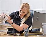 A businesswoman is seated at a desk in an office and is looking through a stack of paperwork.  Horizontally framed shot.