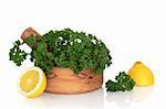 Parsley herb leaf sprigs in an olive wood mortar with pestle and two lemon halves, over white background.