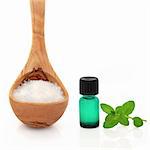 Sea salt in an olive wood ladle with an aromatherapy essential oil glass bottle and peppermint herb leaf sprig, over white background.