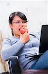 Portrait of a young Asian woman eating apple while looking at her laptop on sofa.