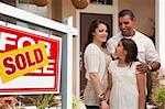 Hispanic Mother, Father and Daughter in Front of Their New Home with Sold Home For Sale Real Estate Sign.