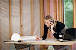 A young woman is looking at blueprints and talking on the phone.  She is working at a construction site.  Horizontally framed shot.