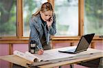 A young woman is looking at a laptop and talking on the phone.  She is working at a construction site.  Horizontally framed shot.