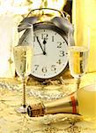 Celebration of the New Year with two glasses of champagne and bottle in the front and blurred alarm clock showing 12 PM in the back. Golden touch