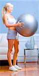 Side view of young woman holding an exercise ball in the living room. A sofa and side table are in the background.  Vertical shot.