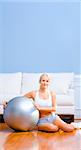 Young woman in sportswear sits next to an exercise ball in her living room.  Vertical shot.