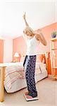 Low angle view of excited woman standing in her bedroom and looking down at a scale. Vertical format.