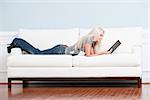 Full length view of woman lying on white couch and reading a book. Horizontal format.