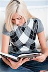 Cropped close-up of woman sitting cross-legged on white couch and reading a book. Vertical format.