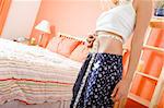 Cropped tilt view of woman standing next to her bed and using a tape measure to measure her waist. Horizontal format.