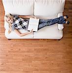 Full length overhead view of woman reclining on white couch with a book, as she looks up at the camera. Square format.