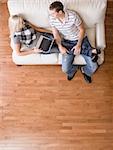 Full length overhead view of couple relaxing together on white couch, with woman using laptop and stretching out with her legs in the man's lap. Vertical format.