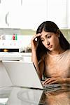 A young woman uses a laptop on a kitchen table. She has a worried expression on her face. Vertical shot.
