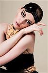 Attractive young woman wearing a black and gold dress with a black fabric hairdressing. She is leaning her head on the back of her hand. Vertical shot.