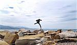 An excited adult full of energy and freedom jumping high over rocks by the sea and a blue sky